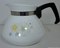 Side view of white ceramic Corning Ware coffee pot with floral bouquet pattern, black handle, silver metal band, scratches visible.
