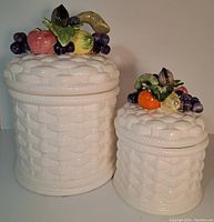 Front view of two white ceramic canisters with basket weave pattern. Lids topped with colorful ceramic fruit including grapes, oranges, bananas, and apples. Larger canister on left, smaller on right.