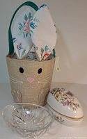 Photo showing burlap woven bunny basket with black stitched eyes and pink nose, containing two floral cloth napkins folded into bunny ears, alongside clear crystal egg-shaped candy dish with cut glass pattern.