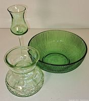 Three vintage green glass items on white surface: candy bowl, candle holder, bulb forcing vase.