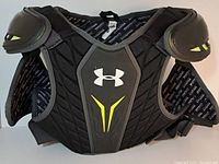 Front view of Under Armour youth lacrosse shoulder pads showing black Coolmax material with yellow and gray accents, chest and shoulder padding with Under Armour logo.