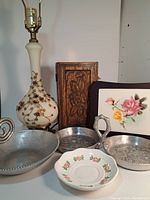 White lamp featuring gold-tone rose metal decorations, wood jewelry box with carved rose detail, small round plate with rose print, two round tin dishes with rose etching, and carving board with rose motifs arranged on shelf.