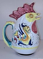 Front side of ceramic rooster-shaped beverage pitcher showing colorful hand-painted decorations, comb, and tail-shaped handle.