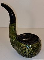 Front and side view of green and black ceramic pipe-shaped ashtray showing embossed designs and black mouthpiece.
