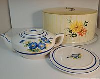 Full view of the ceramic teapot with matching serving plate in front of the cake tin carrier that has yellow orange flowers painted and clasped closure.