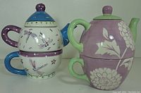 Two ceramic tea for one sets stacked with lids, one floral lavender and green, one dragonfly design with blue and purple accents