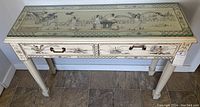 Full front view of the cream wood console table with painted scenic and shell details and two drawers with handles.