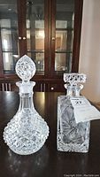 Two crystal decanters displayed on a wood table in front of a glass cabinet, showing their shape and cut patterns.