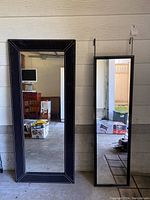Two mirrors standing side by side; left is large with a black stitched frame, right is smaller with over-door hooks