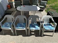 Four white plastic outdoor chairs with blue and white floral cushions arranged in a line on a concrete patio, with patio table and other outdoor furniture visible in the background.