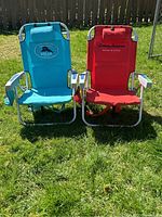 Photo shows two Tommy Bahama outdoor folding beach chairs placed on grass, one turquoise and one red, with logos visible and armrests padded.