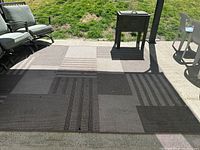 Outdoor rug laid on patio with surrounding chairs and table; shows geometric pattern with various shades of brown and beige squares and stripes.