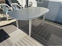 Oval plastic outdoor table shown from an angle on an outdoor mat in daylight. Light grey color with a slatted top and four straight legs.