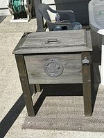 Wooden outdoor cooler viewed from the front showing hinged lid with handle, circular decorative emblem on front panel, four legs, and bottle opener on side.