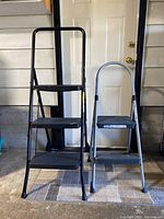 Both step ladders standing side by side showing size and design difference in front of a white door in a garage