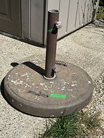 Heavy metal outdoor umbrella stand with circular base, central pole, tightening knob, locking bolt, and two metal handles. Base shows paint wear and chipping. Item placed on concrete with some grass nearby.