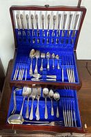 Overall view of the vintage flatware and serving pieces displayed inside a wooden chest lined with blue felt, showing a mixture of knives, forks, spoons, and serving utensils.