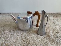 Coffee pot with rounded body, wooden handle and pointed spout, next to a slender metal pitcher with flared top and metal handle on a carpeted floor.