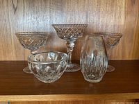 Five pieces of clear crystal glassware arranged on a wooden surface including three stemmed bowls, one small bowl, and one vase.
