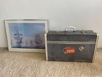 Suitcase next to framed nautical artwork on carpeted floor, showing suitcase front and details of stamps and worn surface