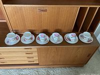 Six teacups and their matching saucers arranged in a row on a wooden cabinet, showing full set.