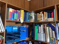 Wide view of two wooden bookshelves filled with books, file holders, blue plastic trays, and blue file boxes. Includes crayons and magazines.