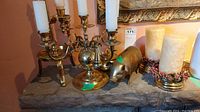 Brass candelabra with five taper candle holders, brass piggy bank, and two brass finish single candle holders with pillar candles on a stone surface.