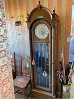 Full view of the grandfather clock in a room with patterned wallpaper, showing its wooden body, glass front door, and clock face.