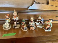 Wide view of six Hummel figurines on wooden surface. Various children characters in traditional clothing presented. One shows girl feeding two geese. Sizes range from 3 to 5.5 inches.