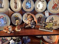 Four Hummel figurines on a wooden shelf with collectible Christmas plates in background. From left to right: cow lying down, photographer with vintage camera, girl holding umbrella, stagecoach with man blowing bugle and horse.
