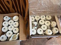 Two cardboard boxes containing multiple white ceramic mugs and creamers decorated with holly and ivy design