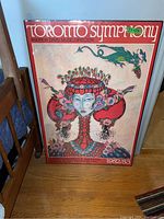 Front view of one Toronto Symphony poster leaning against a chair, showing full artwork with central floral figure and dragon, framed by a red border with text.