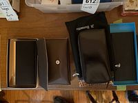 Three black leather wallets on a wooden surface, showing the Birks with embossed lion logo bottom corner, JI Bussetto zip-around wallet with pouch and box, and Renwick wallet with snap closure and box.