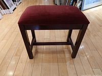 Front view of the Bombay ottoman showing its velvet cushion and wooden legs on light wood floor.