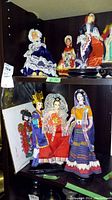 Shelf showing assorted ethnic dolls including a female doll in blue lace dress bonnet, other dolls dressed in various regional costumes including a woman in colorful skirt and orange top, a male in blue and red costume with crown, and another woman in red dress with white lace headpiece.