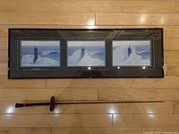 Framed horizontal triptych showing three photos of a lighthouse surrounded by ice and snow, with a rapier sword below it on a wooden floor.