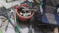 Overview of lot showing long-handled gardening tools, brown pot with smaller tools, parts of leaf blower, and folding chair