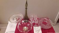 Photo shows tall crystal decanter with stopper, several clear bowls and plates, and a set of six small glasses arranged on a red and white cloth