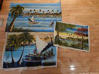 Three canvas paintings laid out on wood floor, showing tropical coastal scenes including boats with sails and trees, as well as a smaller landscape painting.