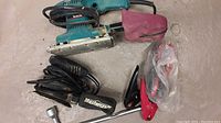 Image showing Makita finishing sander with dust bag and Mastercraft detail sander with power cord. Also red and black jumper cables and L-shaped metal tool are visible.