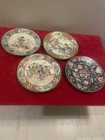 Four vintage Asian hand painted plates on red surface showing front with detailed artwork of traditional scenes and floral designs