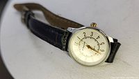 Vintage wristwatch with white dial, gold-tone hands, black Arabic numerals, and black leather strap, angled view