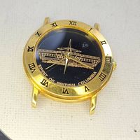 Close-up photo of gold-tone wristwatch featuring a black dial with gold ferry illustration and Roman numeral bezel.
