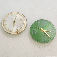 Two Swiss watch movements side by side on white surface. One Rougié automatic with silver and gold finish, date display; the other Ruan A Gogo green dial with large numerals.