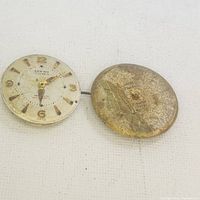 Two vintage watch movements shown side by side on white fabric background, one with dial and hands, the other showing corroded backside.