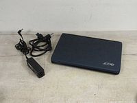 Closed blue ACER Aspire One laptop and separate charging cable with AC adapter on light surface.