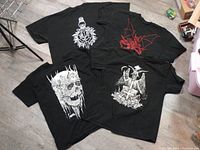 Four black men's graphic tattoo-style T-shirts laid out flat on wood floor, showing front and back designs.
