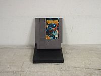 Front view of gray NES cartridge with colorful label showing game title and boxing artwork, positioned on black stand against plain background.