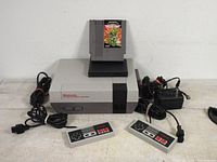 Nintendo NES console with two controllers, power adapter, and game cartridge displayed on top.