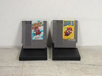 Front view of two original Nintendo NES game cartridges: Super Mario Bros 2 and Super Mario Bros 3, displayed standing upright on black stands.